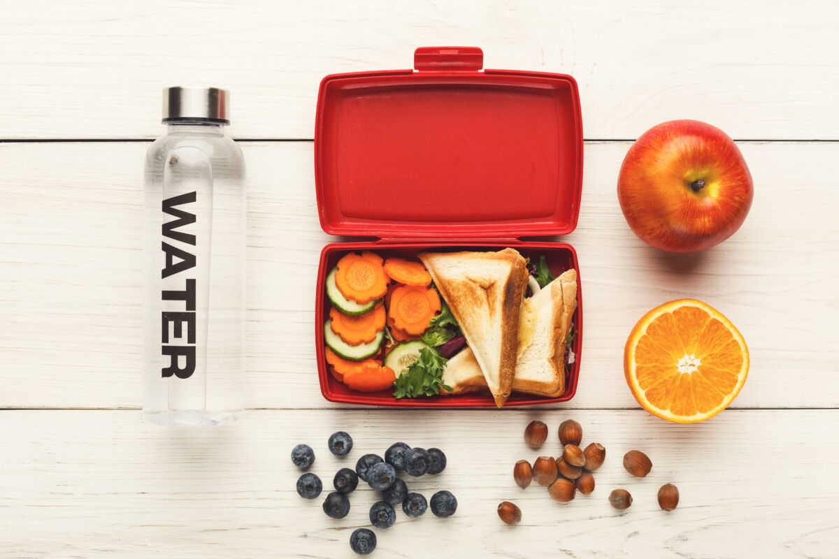 Lunch box filled with mixed vegetables, sandwich, blueberries, nuts and water bottle on white wooden table background